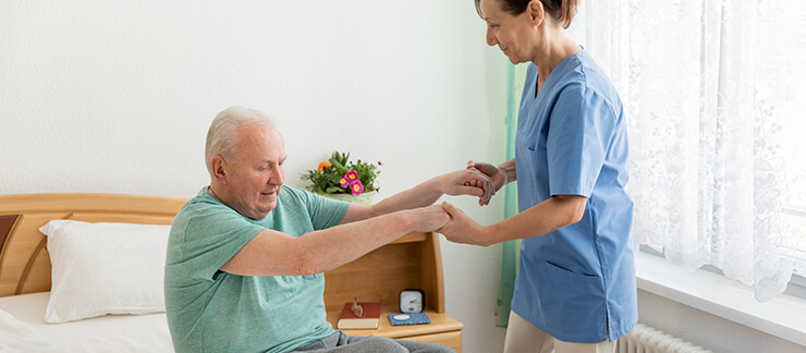 An in-home Alzheimer's care provider stands as she grabs a sitting elderly man by the hands to assist him out of bed
