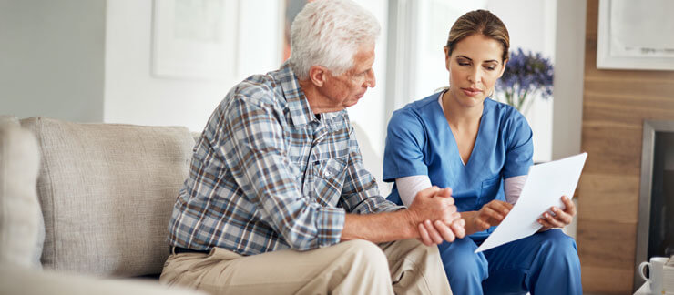 Home care worker with paper in hand discusses care plan with senior male sitting on couch.