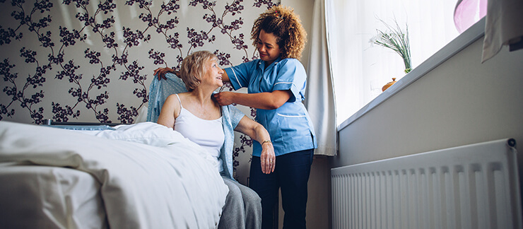 Female care provider helps senior woman in bed to get dressed.