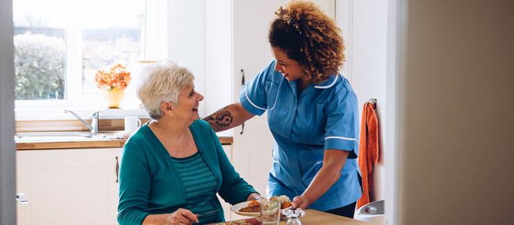 Private duty caregiver serves lunch to senior woman sitting at kitchen table.