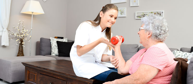 Female caregiver helps elderly woman sitting down at home doing arm exercises with a hand weight.