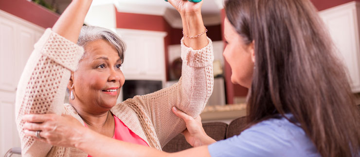 Home care worker helps senior woman raise arms for a sitting exercise.