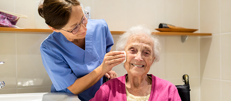 Female care aide helps wash the face of an elderly woman sitting in wheelchair in the bathroom.