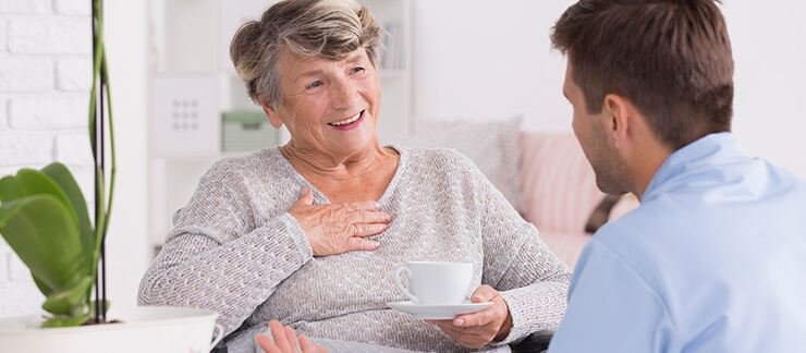 Elderly woman with dementia smiles with coffee cup in hand talking to a male care worker.