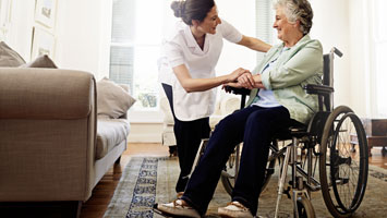 Female care worker holds hand of smiling elderly woman sitting in a wheelchair at home.