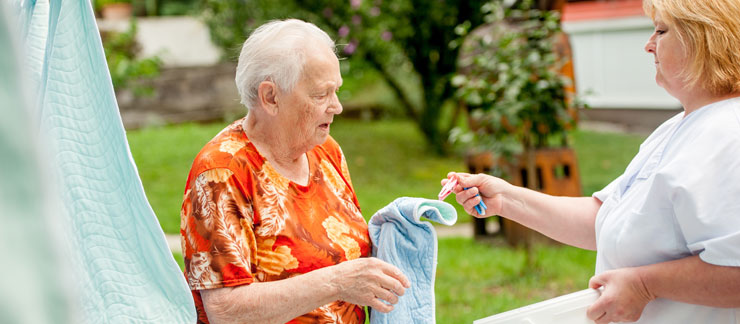 Female home care worker helps senior woman hanging up blanket on clothes line outside.