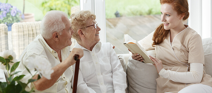 Elderly man with cane sits with his wife on couch listening to a female care provider read to them from a book.