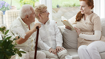 Senior couple listens to a female home care worker reading to them from a book while sitting on the couch together.