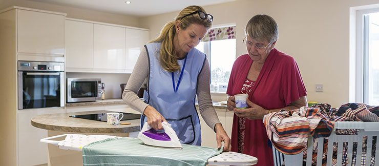 A professional 24/7 caregiver does chores, like ironing, for a senior woman at home.