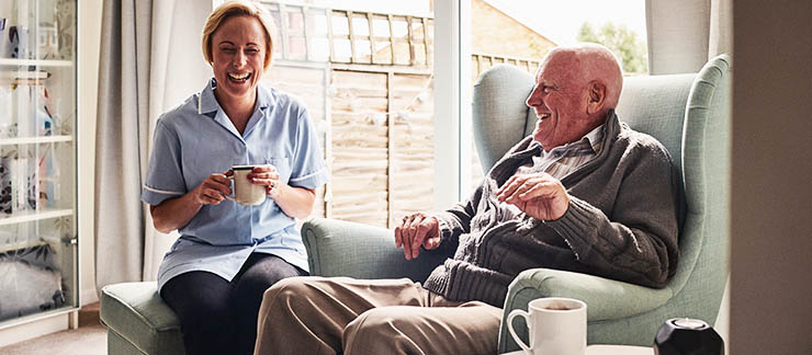 Female companion care worker and senior man laugh while sitting and drinking coffee.