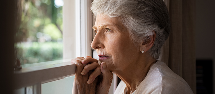 A Sad looking elderly woman stands at the window gazing outside and resting her hands on the window pane