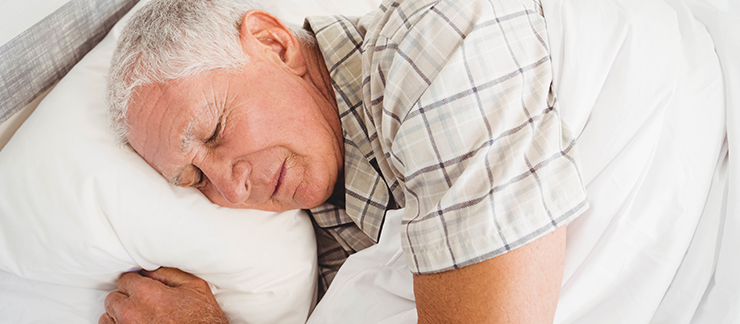 An elderly man with white hair and a plaid collared shirt sleeps on white sheets and a white pillow