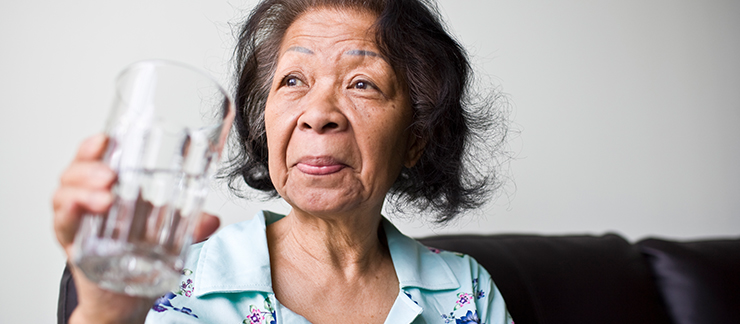 An elderly woman sits on her couch holding a glass of water and gazing into the distance