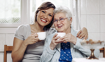 Home care worker visits a senior woman in her own apartment. The female caregiver has arm over the woman's shoulder as they prepare to drink tea.