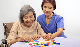 Female caregiver and senior woman playing a wooden shape puzzle game for dementia prevention.