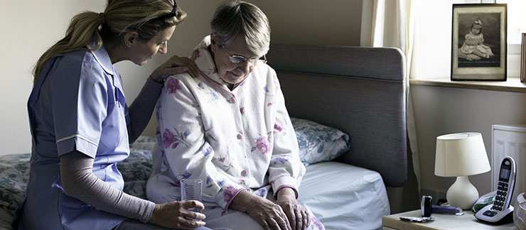 Female caregiver consoles depressed elderly woman sitting on bed with pajamas.