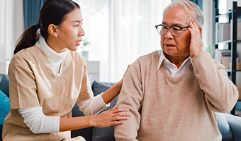 Young female professional caregiver comforts elderly male Alzheimer's patient sitting in living room at home.