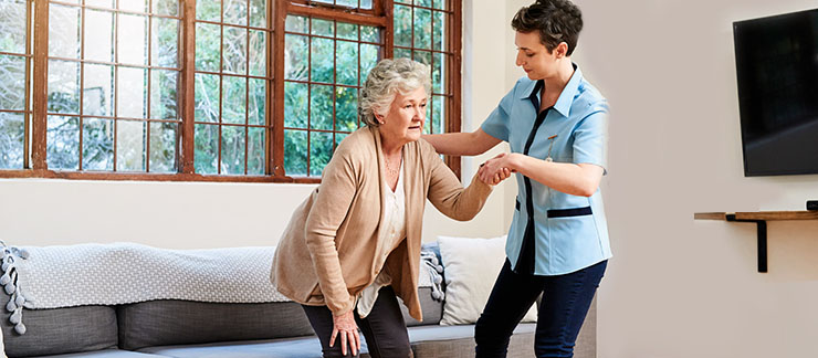 An in-home caregiver helps senior woman with arthritis get off the couch safely.