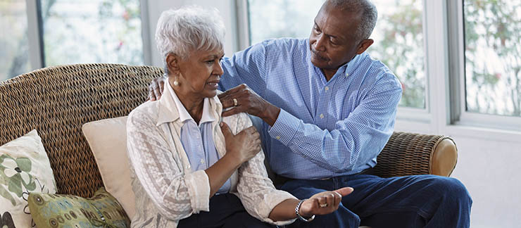 A senior African-American woman sitting at home on a couch, complaining about pain radiating from her shoulder. Her husband tries to provide pain relief by massaging her shoulder.