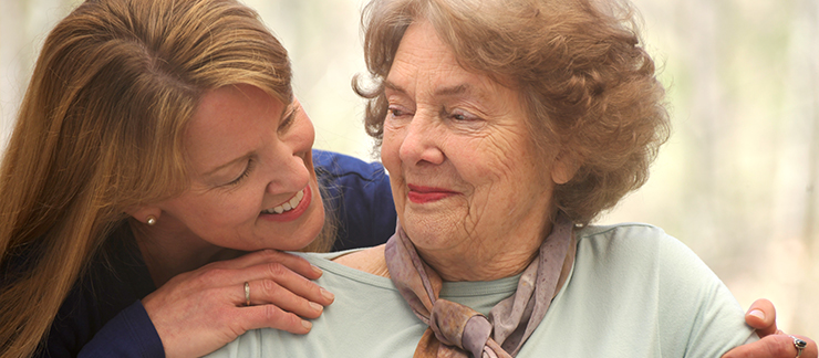 24/7 live-in caregiver smiles while embracing a elderly female client sitting at home.
