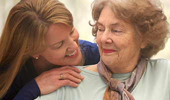 24/7 live-in caregiver smiles while embracing a elderly female client sitting at home.
