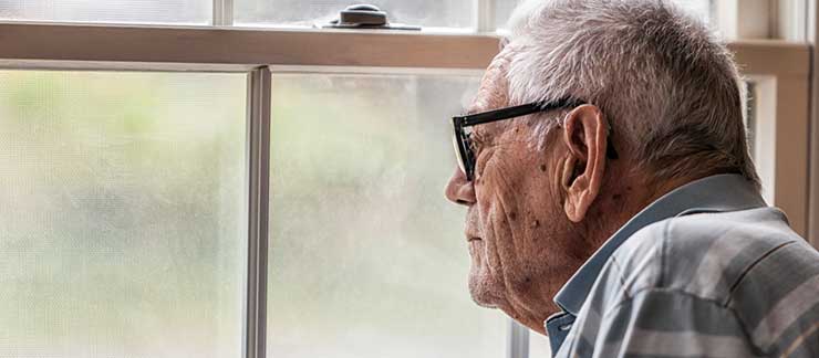 Elderly man looking sad while looking out the window from home.