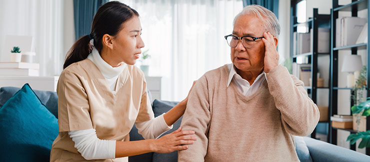 Female caregiver comforts senior man experiencing confusion or brain fog while sitting on couch at home.
