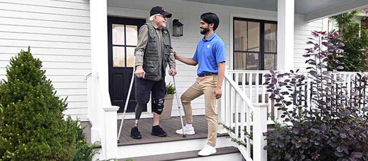 Visiting Angels male caregiver helps Army veteran with prosthetic leg and crutches down the steps of his porch.