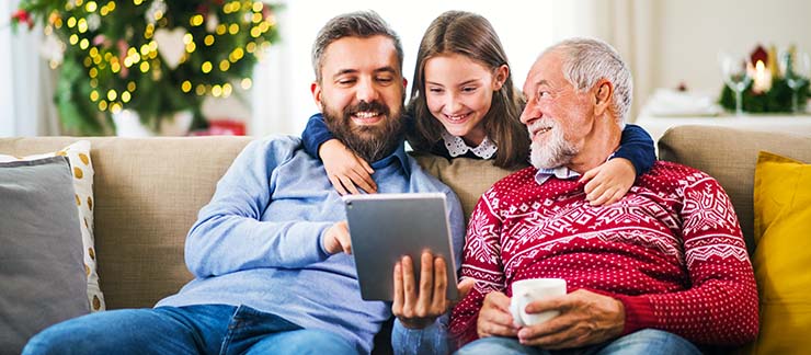 A happy young girl sits on a sofa with her father and grandfather watching a Christmas movie on a computer tablet.