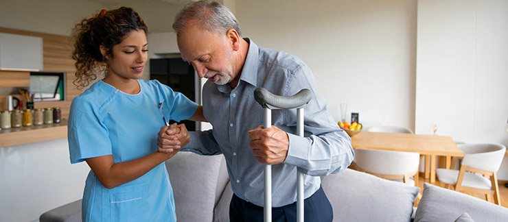 Female in-home caregiver helps senior male with crutches try to walk.