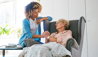 Female caregiver holds hand of elderly woman with back pain as she sits on a chair at home.