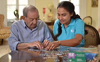 Elderly man  sits at table putting a puzzle together with a helpful Visiting Angels home care worker.