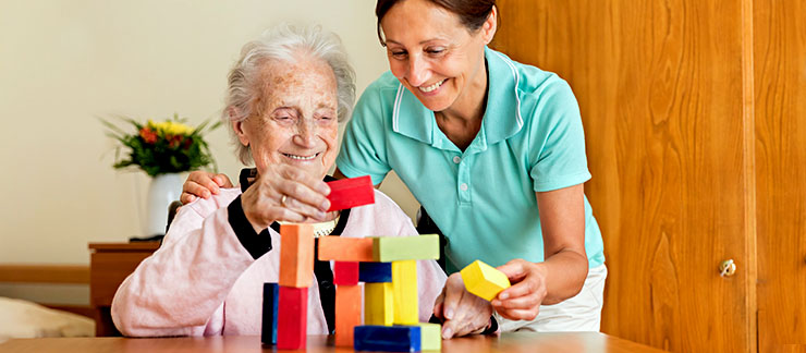 Female caregiver helps smiling elderly woman with building blocks activity at table.