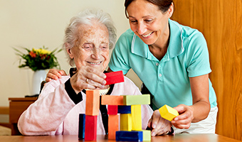Female care worker helps elderly woman build a tower of blocks together on a table.