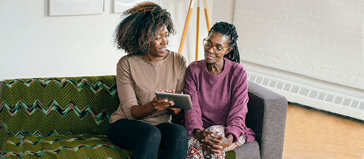 Female caregiver sitting on a couch showing something on  a computer tablet to a senior woman with dementia.