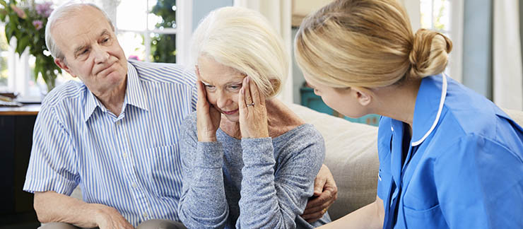 Senior woman with dementia appears upset and is comforted on couch by husband and caregiver.