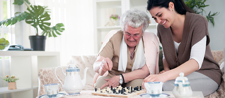 An elderly woman laughs on a sofa at home while playing a game of chess with a female care worker.