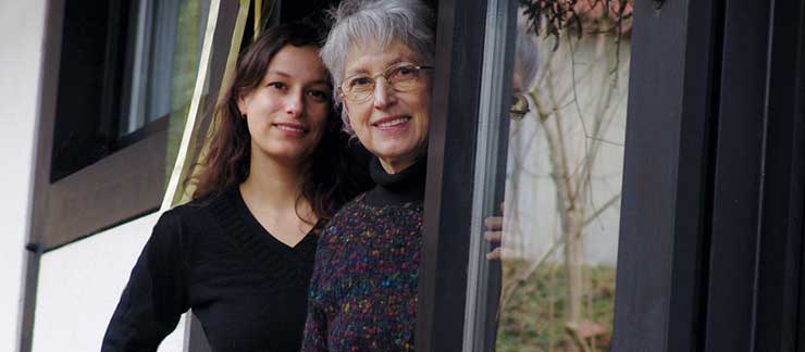 Daughter and senior mother smile while looking outside standing at the front door.