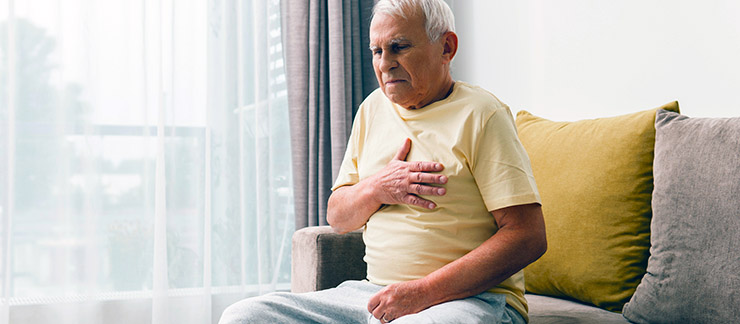 Senior male sitting on a couch at home holds chest after experiencing GERD.