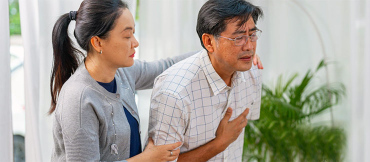 A female caregiver comforts an Asian senior man in his house experiencing chest pain.
