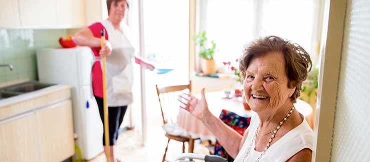 Elderly woman smiles as female care worker with mop cleans the kitchen.