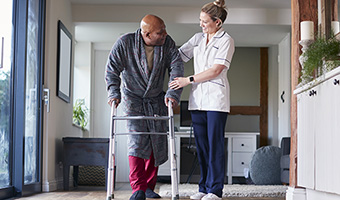 Female care worker helps senior male with a walker down a hallway.