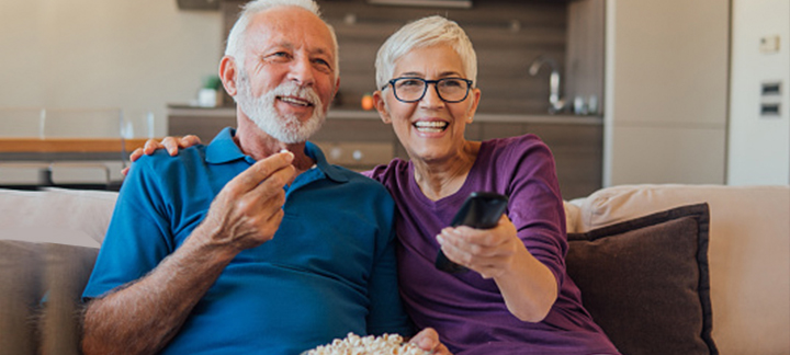 Senior couple smiles while sitting on couch and eating popcorn while watching a Christmas movie.
