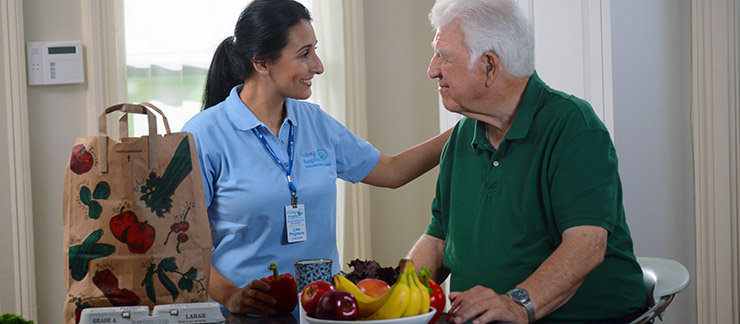 Female Visiting Angels' caregiver greets senior man at home while unpacking groceries after a shopping trip.