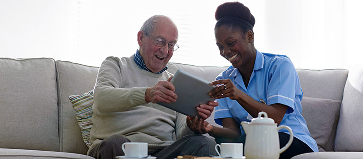 Elderly man on couch smiles while getting assistance with his computer tablet from a helpful home care aide.