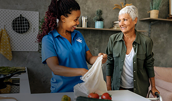 Female caregiver brings groceries to a happy senior woman standing with a cane inside her home.