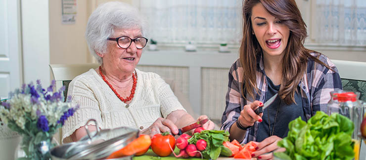 Female caregiver assists eldery woman with cutting vegetables safely at the kitchen table.