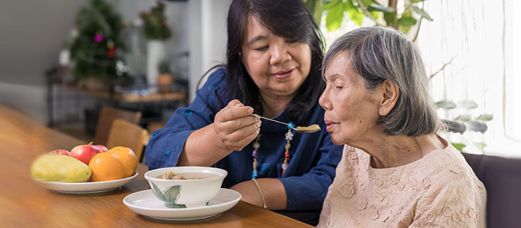 A daughter helps feed a bowl of soup to her elderly mother at the kitchen table.