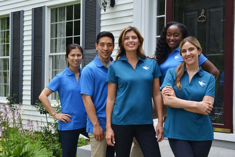 five home care caregivers in blue and teal uniforms standing on a porch smiling 