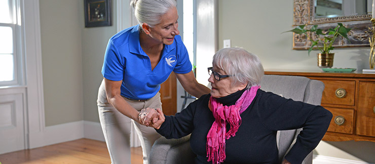 A female Visiting Angels non-medical caregiver helps elderly woman arise from a chair at her home.
